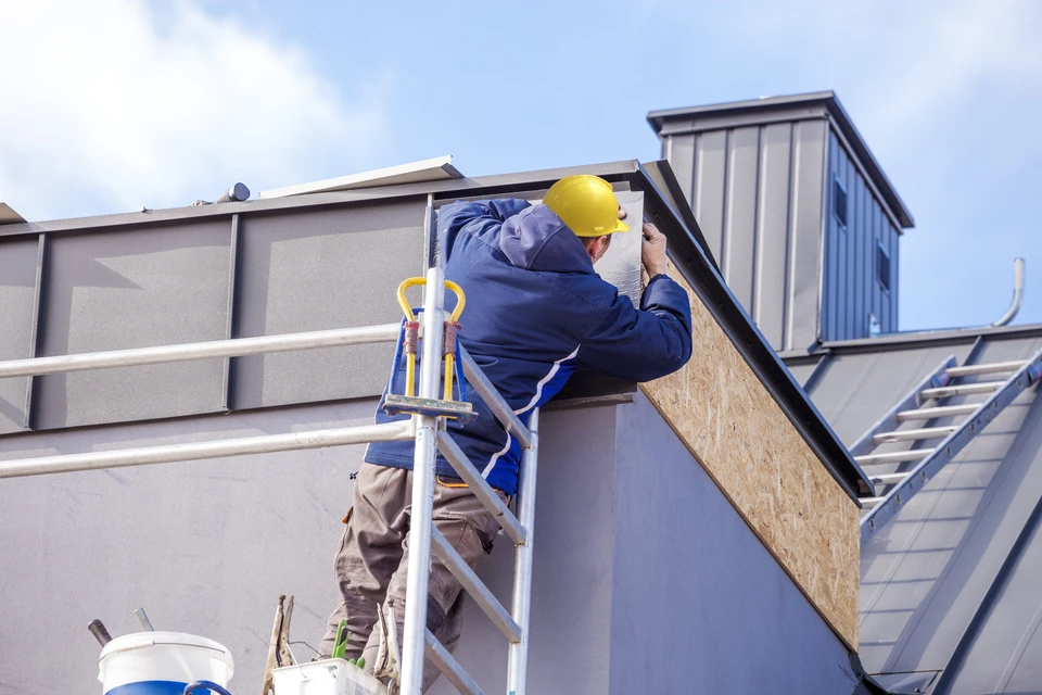 Chantier de construction extérieur avec ouvrier en bleu de travail et casque jaune installant un panneau isolant sur façade depuis échafaudage mobile
