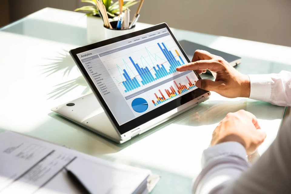 Personne en chemise blanche utilisant un ordinateur portable sur un bureau en verre, pointant du doigt des graphiques analytiques affichés à l'écran